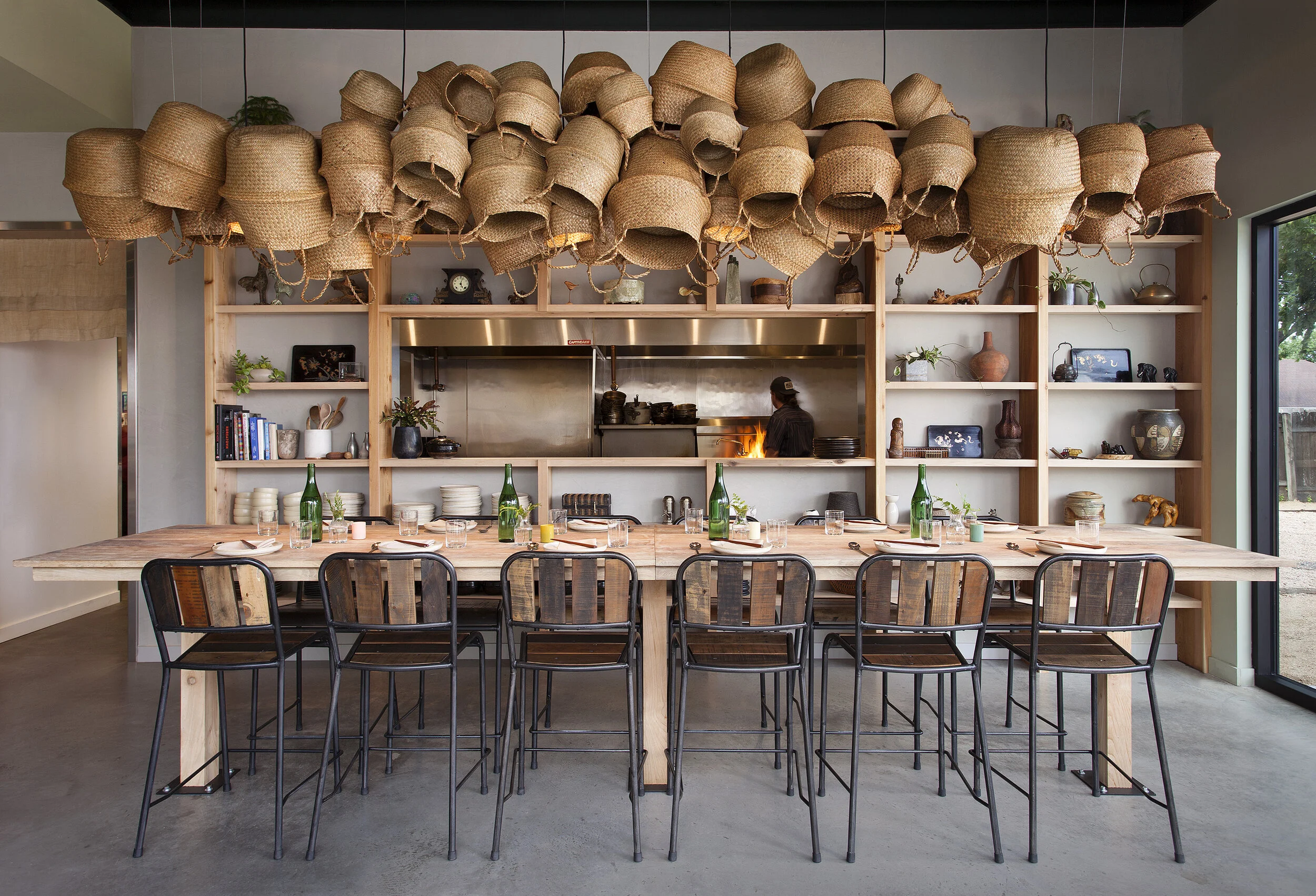 Wood counter with black metal bar stools, woven-back seating, open shelving wall, and hanging rattan pendant lights